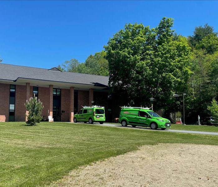 Two green SERVPRO vehicles parked outside large brick buildings.